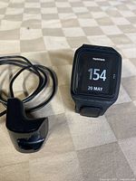 Front view of TomTom watch face showing digital time and date alongside USB charging dock and cable on beige patterned surface.