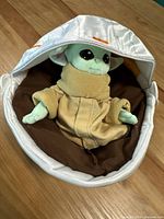 Front view of Grogu plush toy seated inside white and orange carrying pod with brown interior lining