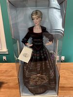 Front view of full porcelain doll in packaging, showing outfit and included tag.