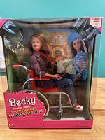 Front view of boxed Becky doll sitting in red wheelchair, wearing plaid jacket, blue jeans, light brown hair, holding camera accessory.