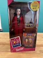 Front view of boxed Rosie O'Donnell doll with red suit and activity book inside packaging.