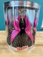 Front view of Barbie doll in original silver packaging with clear front, doll wearing black rhinestone gown and pink shawl, tiara visible, 'Happy Holidays' text at the top, Barbie logo on bottom.