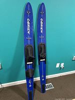 Full view of two blue O'Brien water skis standing upright against a teal wall showing overall condition and length.