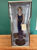 Porcelain doll in original box, showing full figure dressed in dark blue dress with accessories and display plaque.