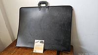 Black artist portfolio with handle placed on wooden surface showing overall condition and size.