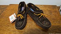 Pair of dark brown suede moccasin-style shoes with fringes, beaded decoration, tan leather lace, and original tag attached, placed on wooden surface.