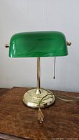 Banker's lamp showing the glossy green glass shade on a polished brass base with visible pull chain and electrical cord unplugged on wooden surface.