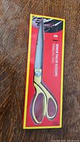 Photo of CMQ scissors in original packaging showing long silver blade and gold-tone handles on a red and yellow box with branding.
