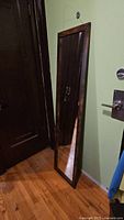 Full view of the tall mirror leaning against a green wall and wooden door, showing dark brown wooden frame and clear glass reflecting the door and metal door handle.