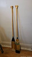 Two Feather Brand wooden canoe paddles leaning against a wall, showing their full length and T-grip handles.