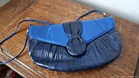 Blue medium-sized handbag with eelskin texture and flap closure featuring a circular detail, placed on wooden surface.