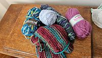 Photo showing six partial skeins of yarn and a knitted swatch sample on a wooden surface. Colors include blue, yellow, burgundy, teal, purple, light blue, and pink.