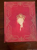 Front cover of one book showing ornate gold decorations on red cloth cover with some wear and fading to the central shield emblem.