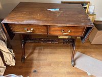 Front view of antique mahogany desk with two drawers, drop-leaf sides down, brass handles, and brass claw feet, showing wear on wood surface.