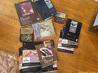 Photo showing multiple pastel paper pads, including Strathmore and Canson brands, used chalk pastels in metal tins, and a clear plastic bag with additional pastel supplies.