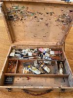 Opened wooden artist box showing paint stained lid and several squeezed oil paint tubes inside compartments.