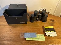 Black Bushnell binoculars next to hard carrying case, lens cloth, and manuals on wooden surface.