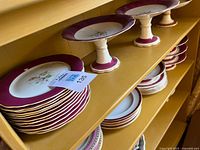 Six ceramic pedestal base plates with burgundy red trim on a yellow shelf.