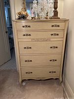 Front view of French style tall dresser showing five drawers, ornate metal handles and carved detailing on bottom drawer and base.