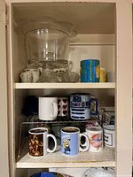 Top shelf showing a clear glass cake stand and assorted mugs including a colorful one, small floral cup, and blue and white cups.