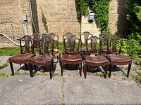 Wide view of nine chairs arranged on an outdoor surface showing carved wood shieldback design and brown leather seats.