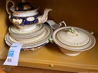 Photo showing an antique teapot with blue and gold decoration stacked above matching plates, alongside a covered bowl with lid and a gravy boat in cream and green patterns on a wooden shelf.