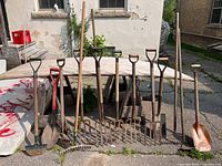 Full view of 12 garden tools including pitchforks, rakes, and shovels arranged in a row outdoors on pavement.