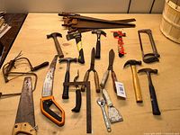 Photo showing full lot of various hand tools laid on a wooden surface including saws, hammers, axe, and other small tools.