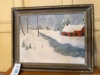 Framed oil painting showing winter snow scene with cabin and river