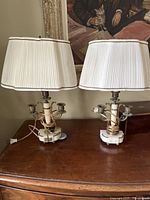 Pair of antique lamps with marble bases, pleated fabric shades, and candle holders on wooden surface in front of a painting.