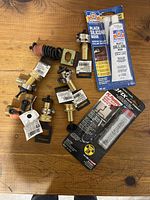 Top-down view of various brass and metal hose fittings, clamps, and two tubes of black silicone sealant on wood surface.