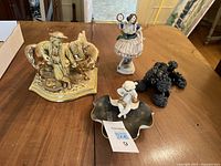 Lot of three porcelain figurines on wooden table including a golden horse with a man, a girl figurine, and a black decorative dish with a white cherub.