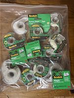 Plastic bag containing multiple Scotch Magic Tape rolls with dispensers and some refill rolls in original packaging.