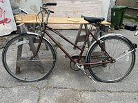 Side view of vintage 3-speed bicycle showing brown frame, fenders, rear rack, flat tires, chain guard and rusty spots on metal parts.