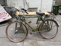 Full side view of bicycle showing olive green frame, fenders, wheels and rear rack against wooden stand