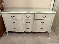 Front view of white dresser showing 9 drawers, curved front, and ornate drawer pulls. The glass top has some visible stains and wear spots.