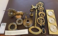 Vintage brass items including unicorn statue, engraved ashtrays, large key with bell, candlestick holder, decorative brass plaques, brass instrument plaques, on a wooden surface with an information note stating they date back to at least 1950s
