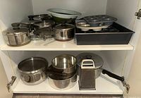 Wide view of metal cooking pots and pans with lids on two shelves showing pots, bowls, baking pans, and muffin tin inside a cabinet.