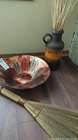 Photo showing ceramic bowl, ceramic pot with wheat decor, and grass broom on floor