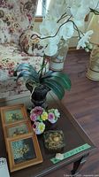 Photo showing faux white orchid plant on table beside three framed floral paintings, vintage china bouquets, and decorative box.