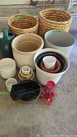 Photo showing two large woven wire baskets, green plastic watering can, multiple ceramic and plastic pots of varying sizes, and a red hummingbird feeder in front