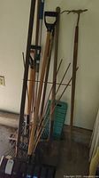Multiple garden and snow tools including rakes, shovels, and brooms grouped together in garage corner