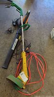 Full view of electric weed wacker with attached orange extension cord and battery powered blower alongside on the ground.
