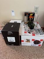 Photo of boxed Moderna 36 oz glass decanter, green glass candleholders, clear tealight holders, and boxed The Un-Candle item on carpet.