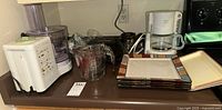 Countertop view showing various kitchen appliances including a large white electric food processor with attached clear plastic bowl, a clear glass measuring cup, a white drip coffee maker with glass carafe, and a set of colorful square ceramic plates stacked.