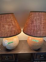 Pair of lamps side by side on a wooden surface, showing their rounded ceramic bases and brown fabric shades with textured pattern.