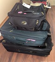 Top view of three stacked American Tourister luggage pieces showing carry-on, medium, and large suitcases in dark fabric with gold zippers and pink zipper pull tabs.
