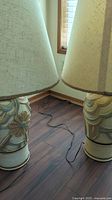 Close-up photo of the two ceramic vase style lamps with beige fabric shades on a wooden floor near a window.