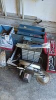 Photo showing handheld circular saw, rectangular toolbox, small parts drawers, and hand tools packed in a garage corner.