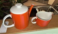 Tea pot and cream jug shown together, highlighting matching vibrant orange color with white accents.